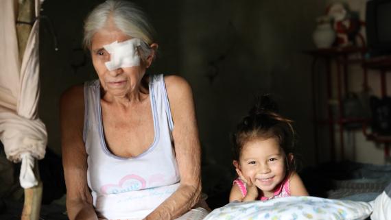 An old woman - with her eye bandaged - sits next to a young girl