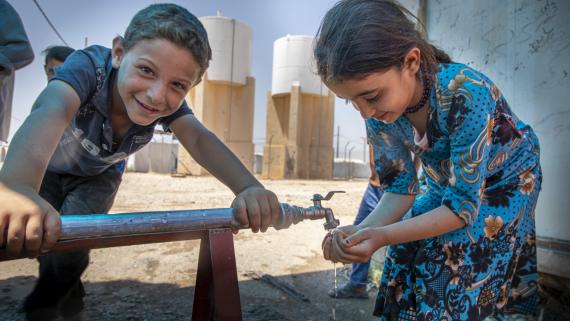 A boy smiles at the camera while a girl washes her hands