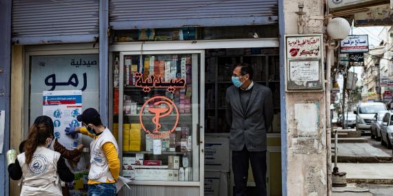 A man stands in front of a pharmacy while two volunteers hang a poster providing instructions on how to protect from COVID-19 